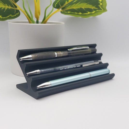 Angled front view of the black pen display holding three pens beside a potted plant.