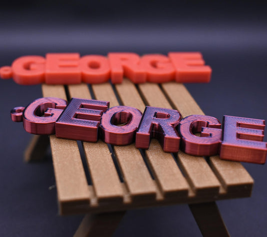 Red and purple George name tag keychains displayed on a miniature wooden bench.